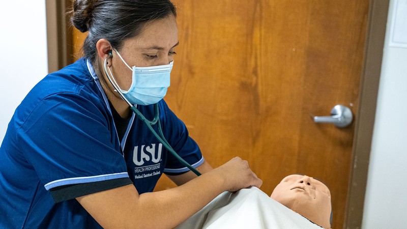 A woman in personal protective equipment uses a stethoscope on a dummy.