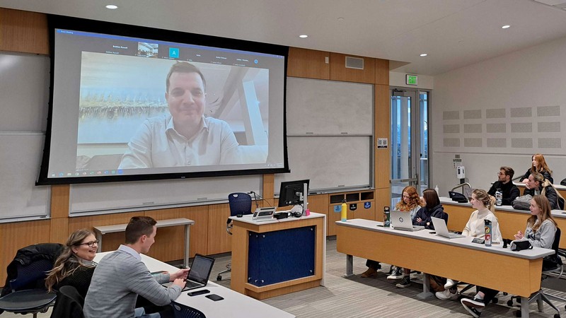 A man speaking to students in a classroom via a video call projected on an overhead screen.