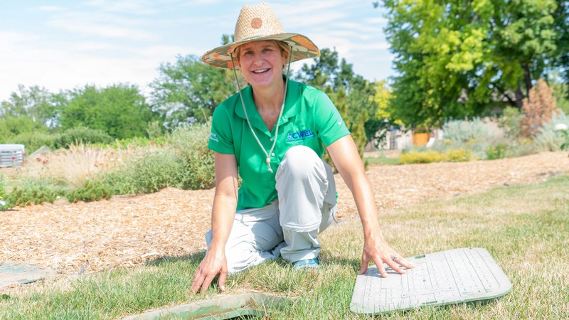 Kelly Kopp kneels by a water valve box on a lawn.