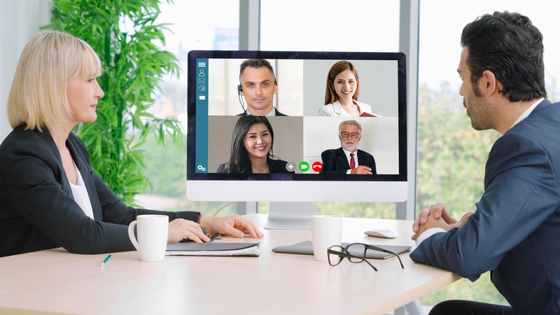 Two people at a desk speak with four others through a computer video conference.