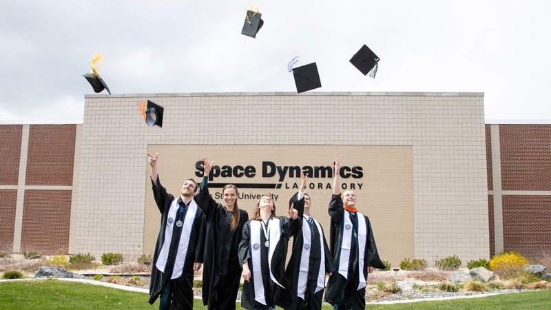 Five students in graduation regalia throw their caps in the air in front of SDL in North Logan.