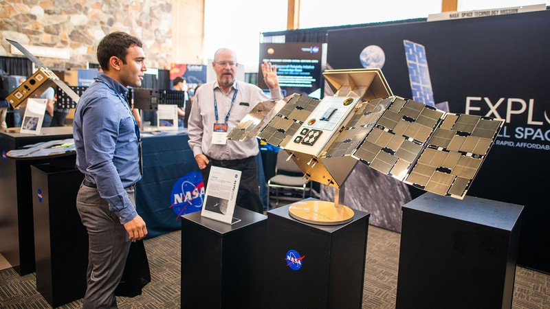 Two people in business attire look at a small satellite demonstration.