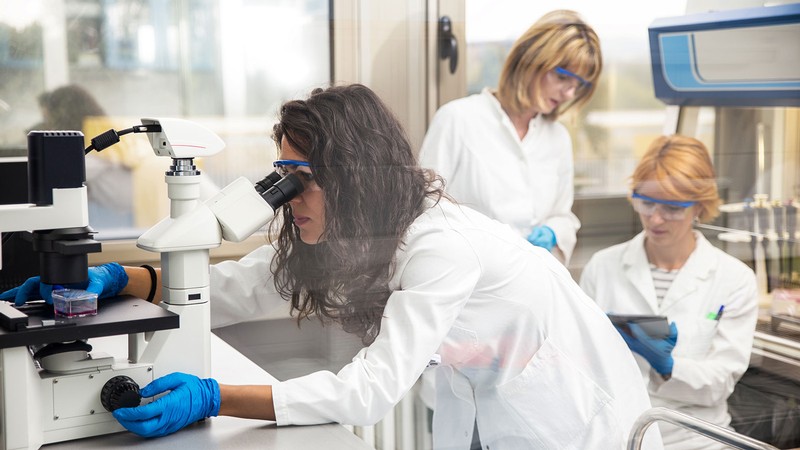 A woman in a lab coat looks into a microscope.