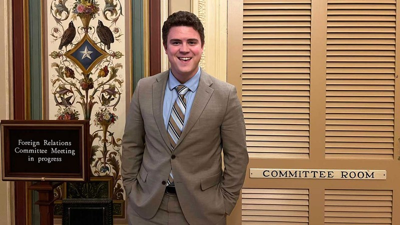 Chase Harward next to a sign that says "Foreign Relations Committee Meeting in progress."
