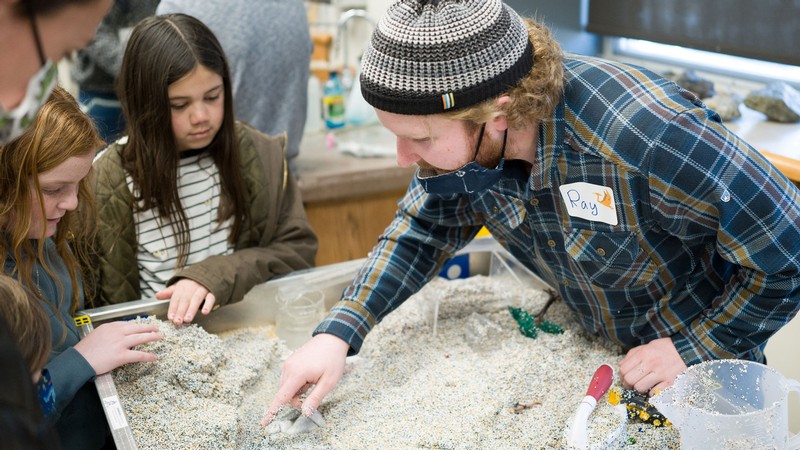 A person digging through a fossil sandbox while children watch.