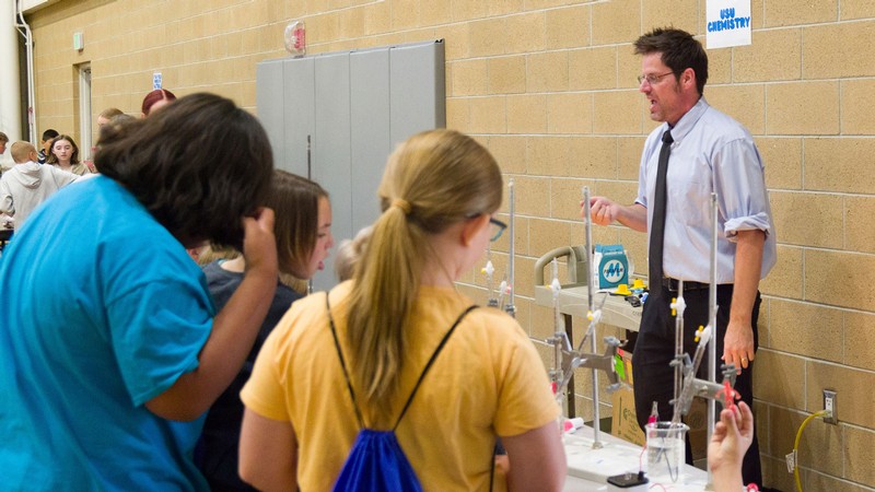 Students watching a chemistry demonstration at a table set up in a school gym.