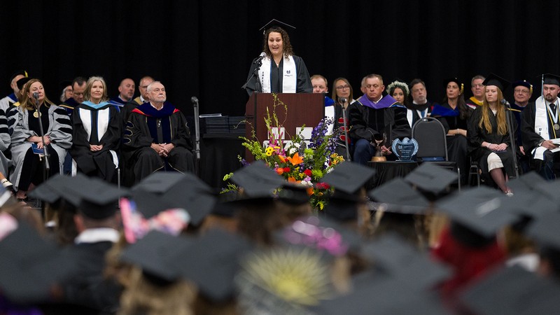 A woman in graduation regalia addresses a crowd.
