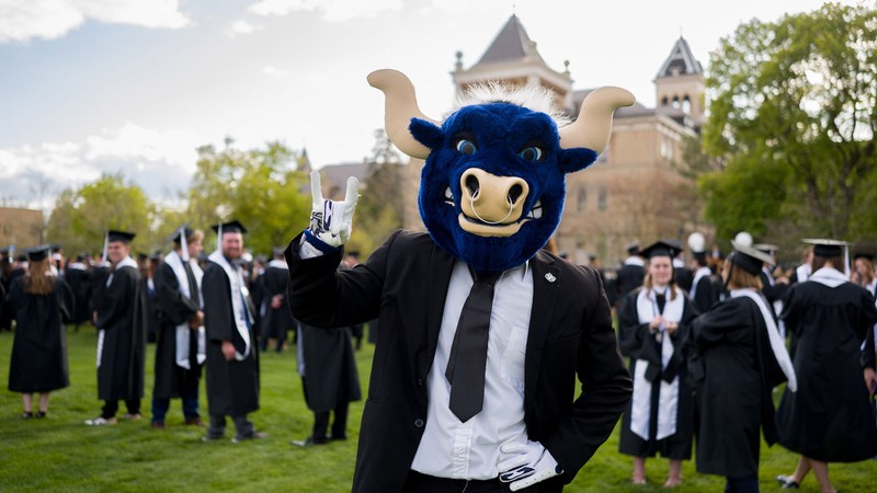 A blue bull mascot wearing a suit and tie poses with people in graduation robes.