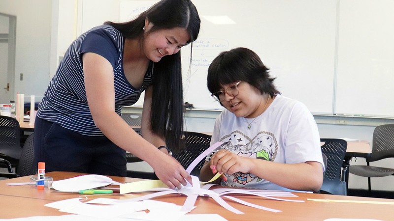 Two women weaving a paper basket.