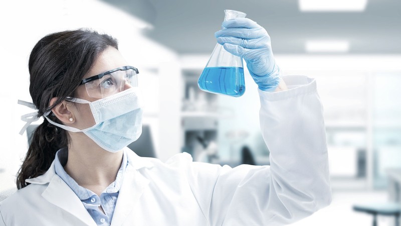 A woman in a lab coat and personal protective equipment holding a flask in a laboratory.