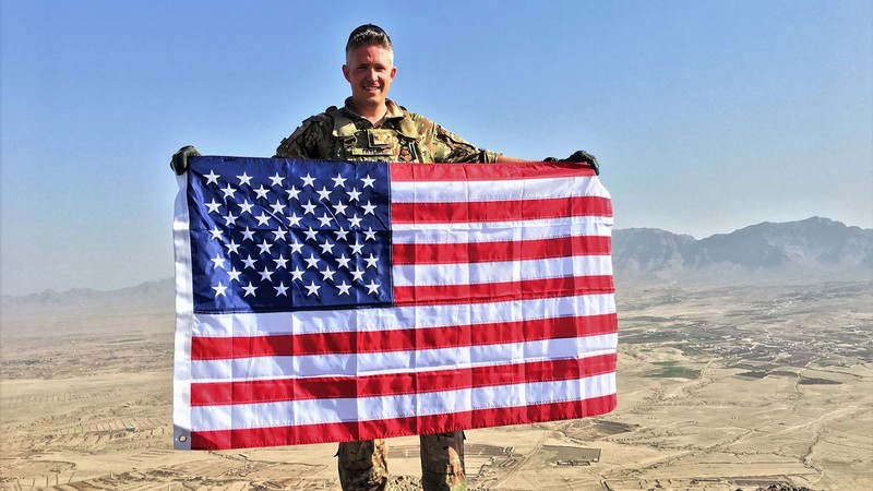 Brent Taylor in Army Fatigues holding a U.S. flag in front of a desert valley.