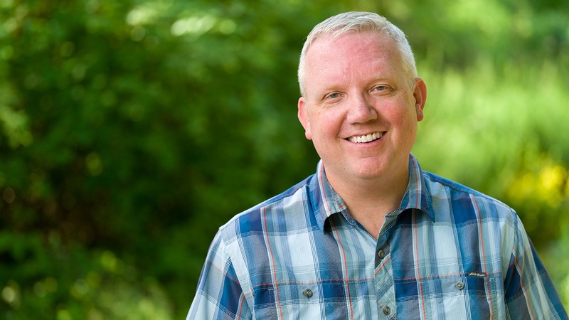 A man poses for a photo in a wooded area.