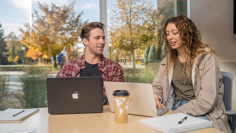 Two students sit at a desk looking at laptops