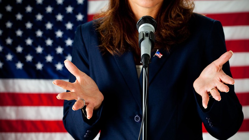 A woman stands at a microphone while wearing a suit and standing in front of an American flag.