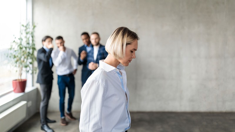 A woman hangs her head as men in the background look at her and laugh to themselves.