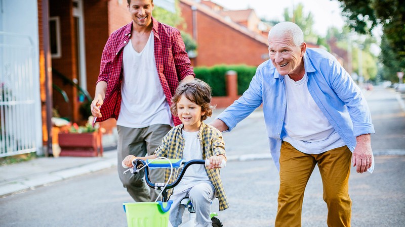 A father and grandfather help a young boy ride a bike.
