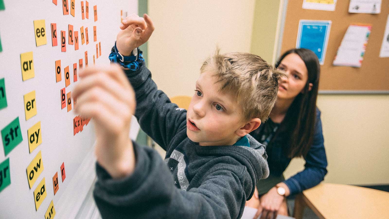 young student moving words and letters around on a bulletin board as his teacher looks on.
