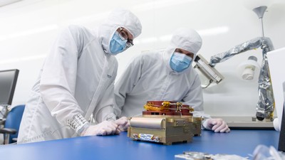 Two scientists in masks and gear work on a camera component.