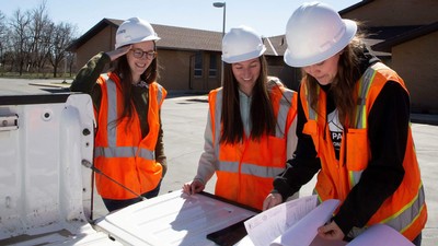 Three students in hard hats review blueprints on a pickup truck tailgate in a church parking lot.