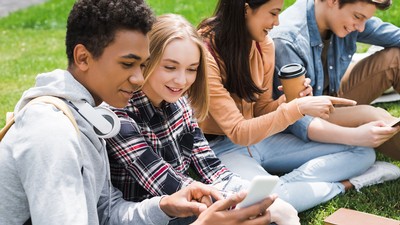 Teens use cell phones while sitting on the grass.
