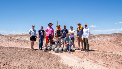 Students posing next to a robotic rover in a desert landscape.
