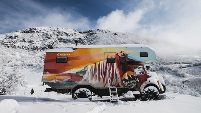 An all-terrain RV with a coastal cliff painted on the side parks in a snowy field.