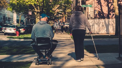 People with disabilities walking down the street, one on a scooter, the other with a cane.