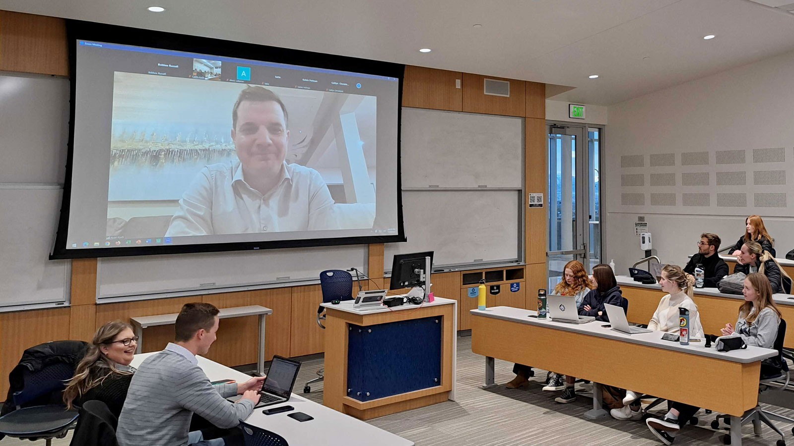 A man speaking to students in a classroom via a video call projected on an overhead screen.
