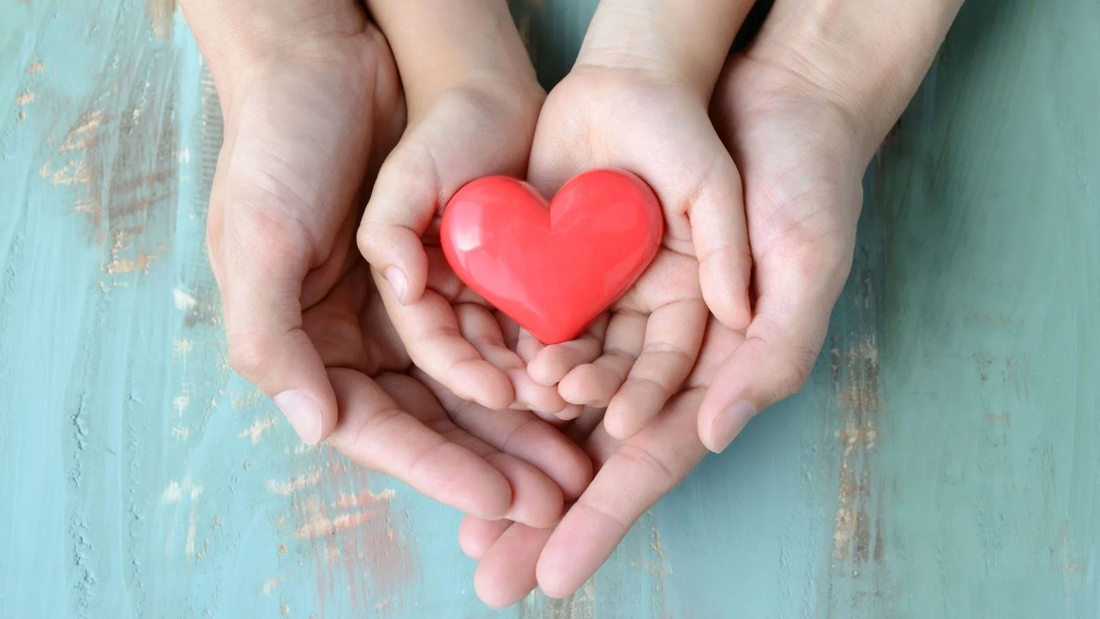 A photo illustration shows a parent's hands holding a child's hands holding a decorative red heart.