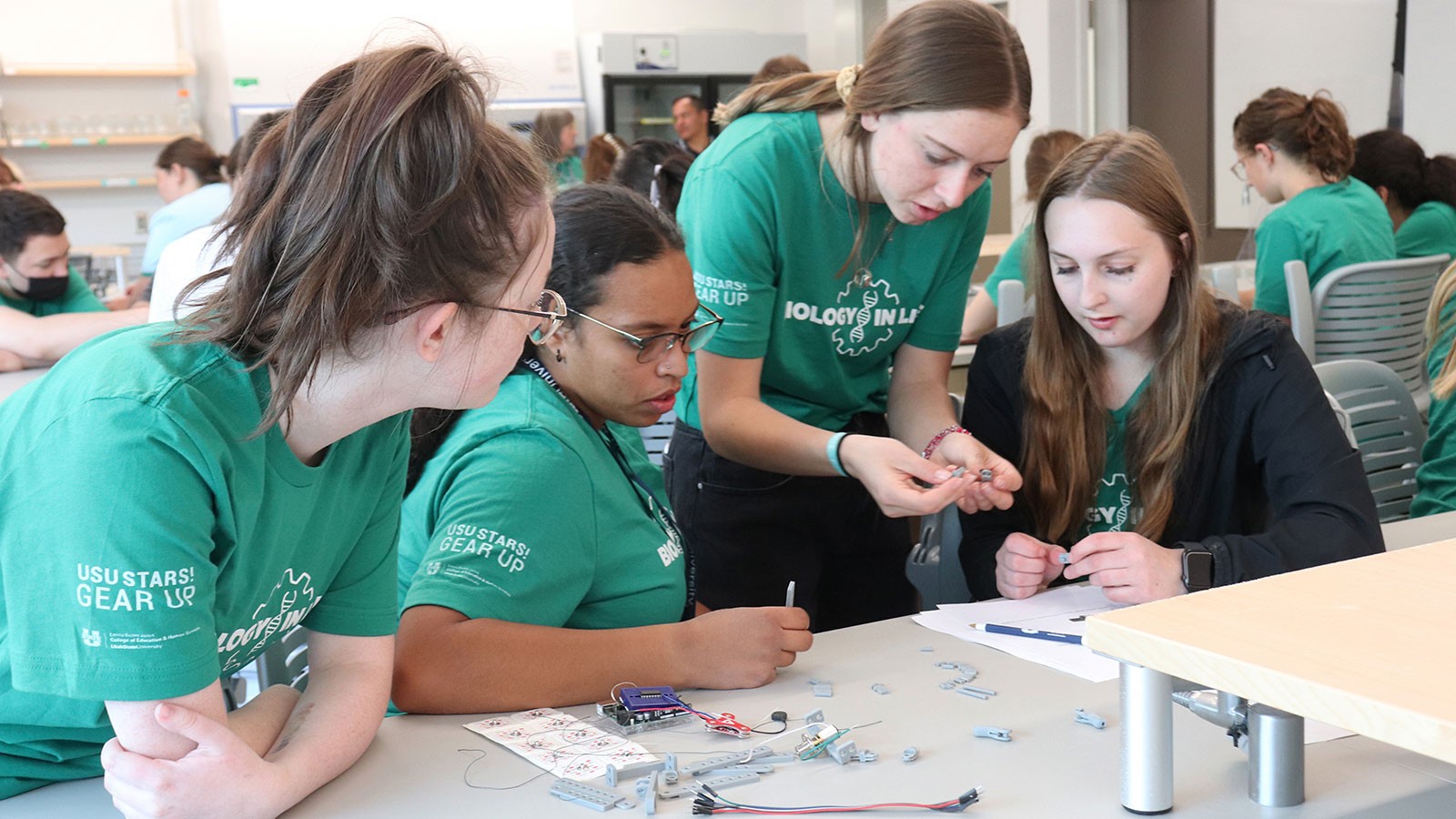 A college student helps three teens build an electronic device in a laboratory classroom.