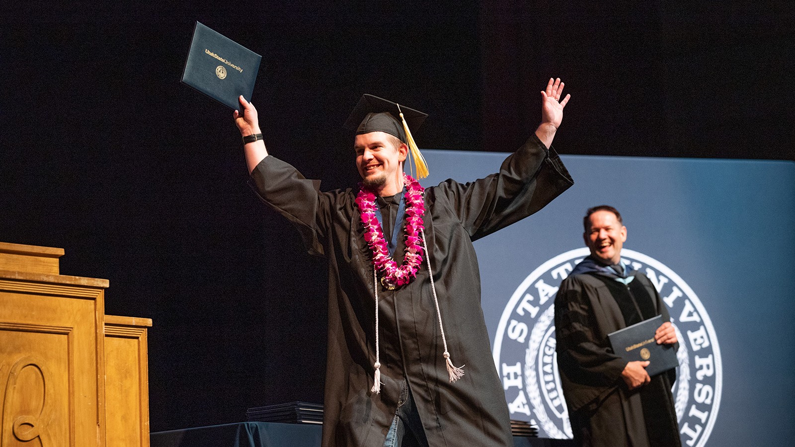 A person wearing graduation regalia and a flower lei holds up a diploma cover.