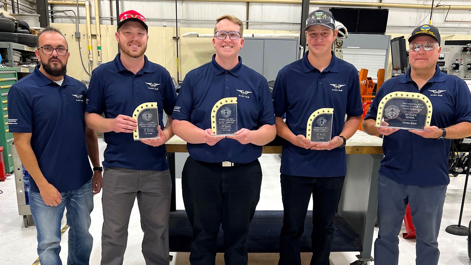 People holding trophies in a mechanic's shop.