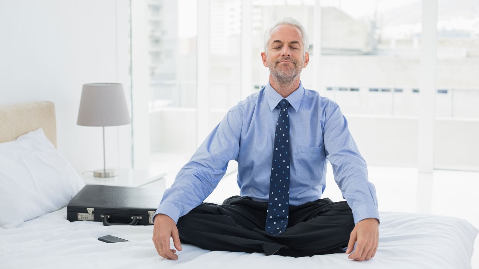 A man in business attire meditating on a hotel bed.