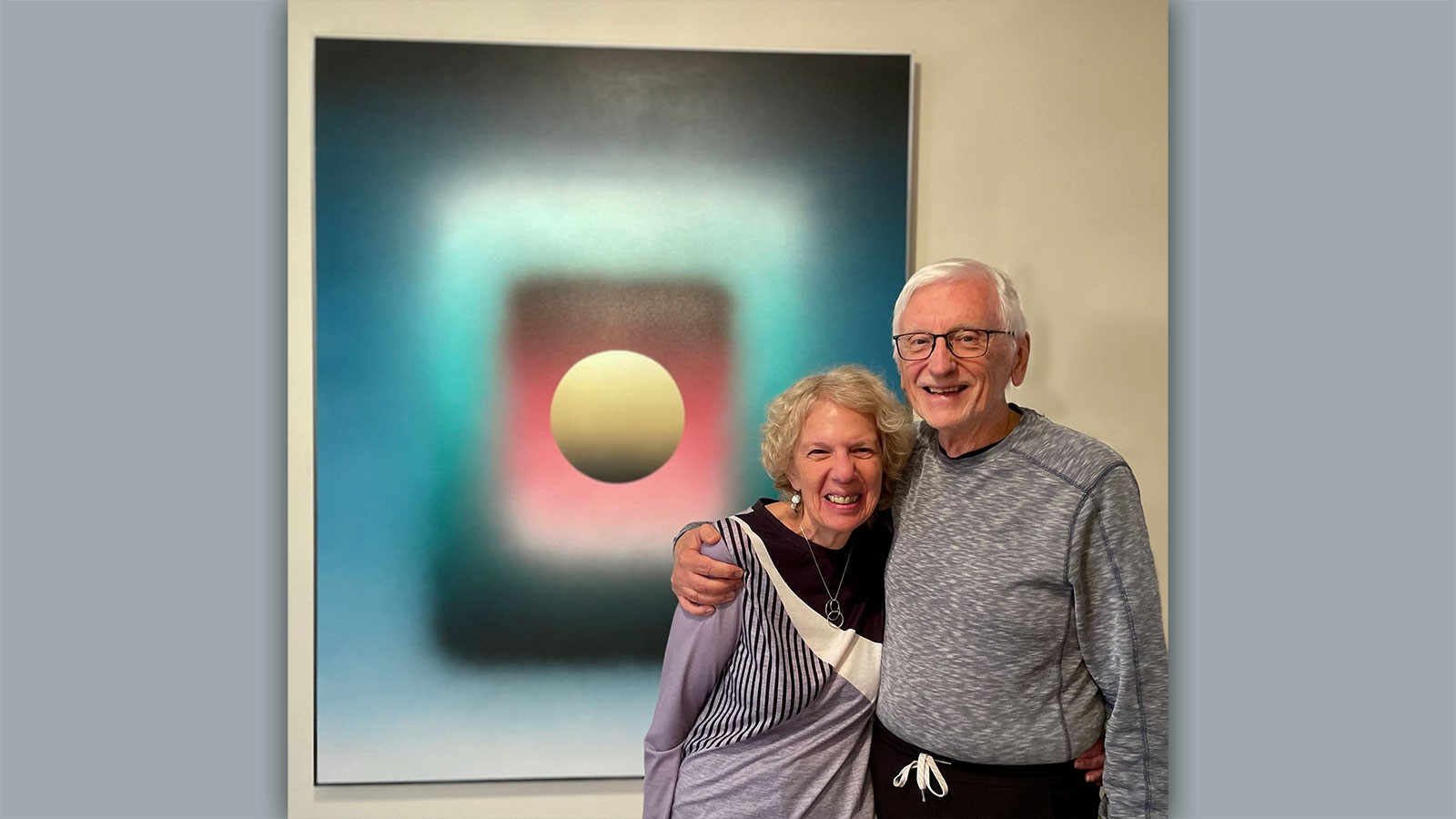 Terry and David Peak standing by an abstract painting with blue, yellow and red geometric shapes.