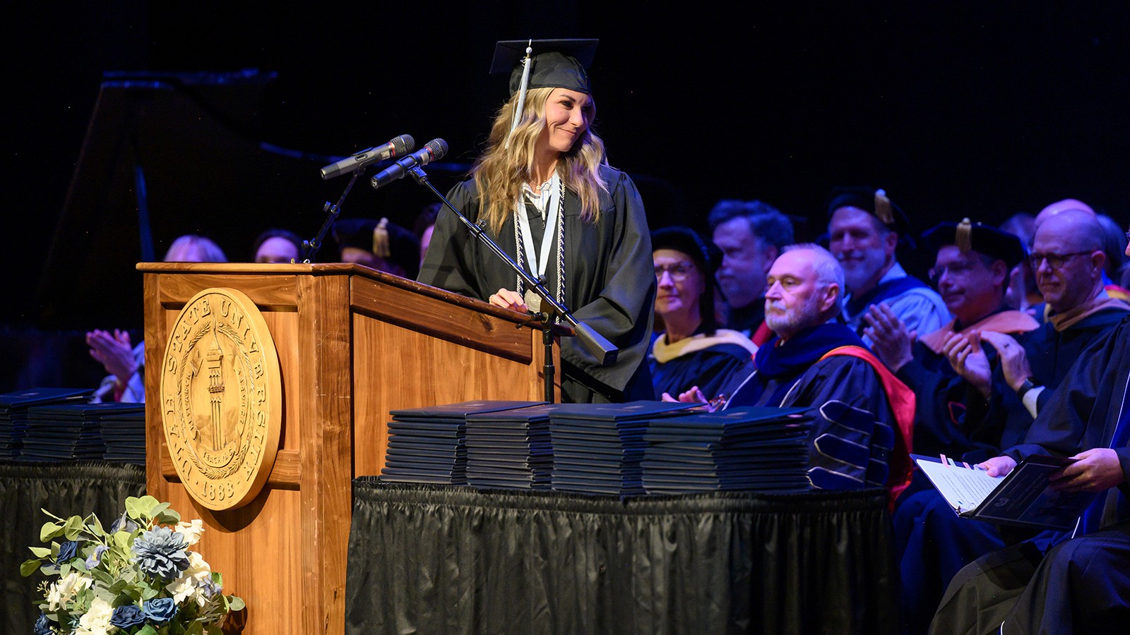 A woman in graduation regalia addresses a crowd.