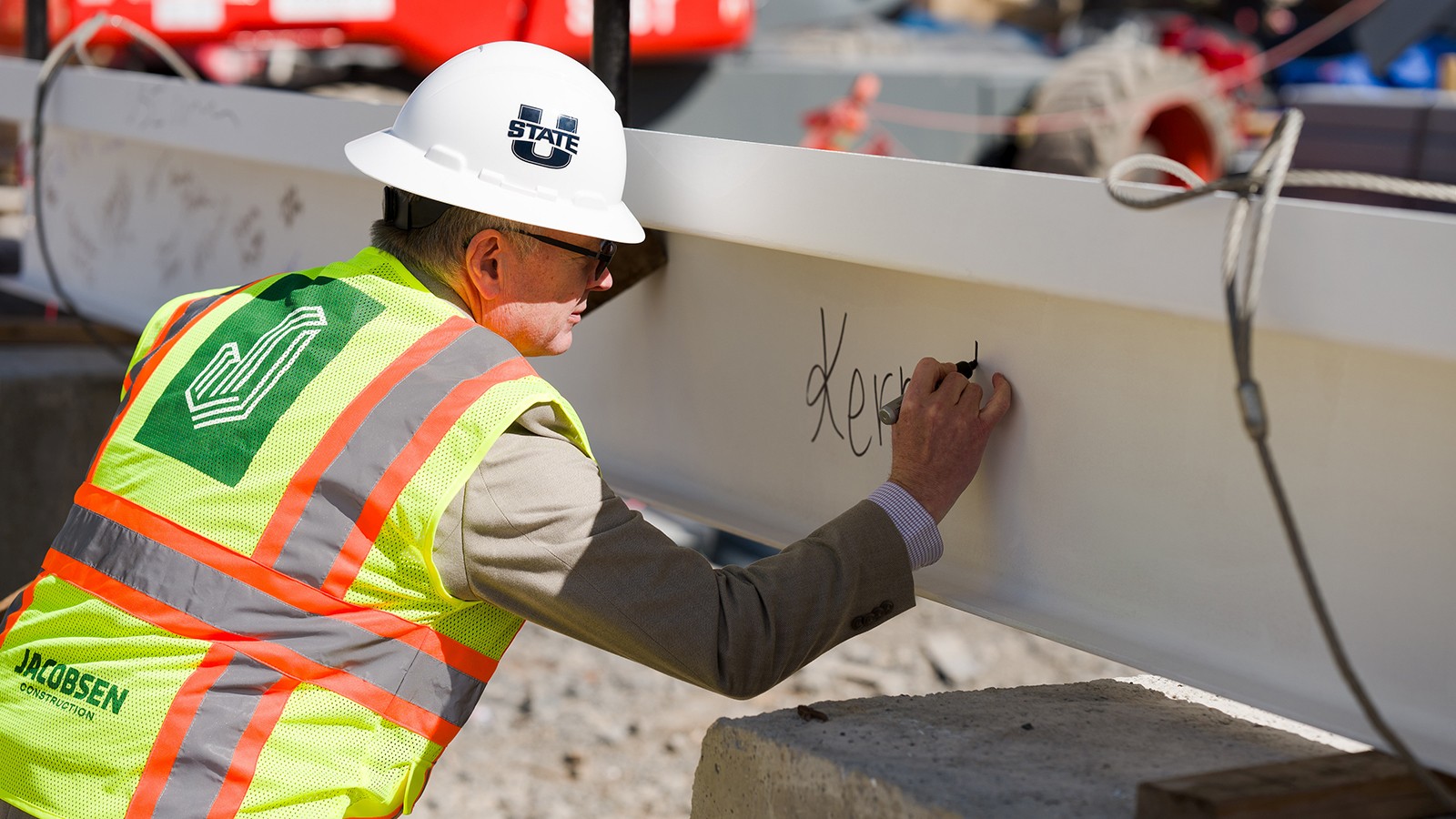 A man in a construction hat and vest signs a steel beam with a marker.