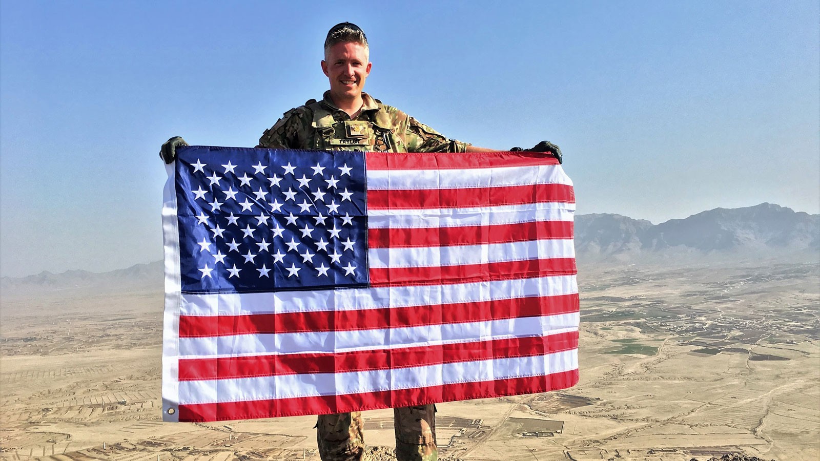 Brent Taylor in Army Fatigues holding a U.S. flag in front of a desert valley.