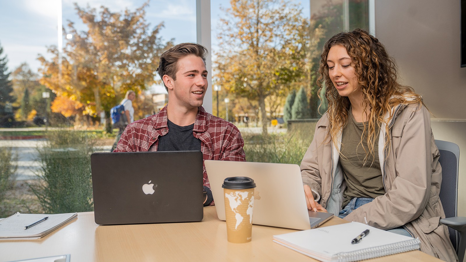 Two students sit at a desk looking at laptops