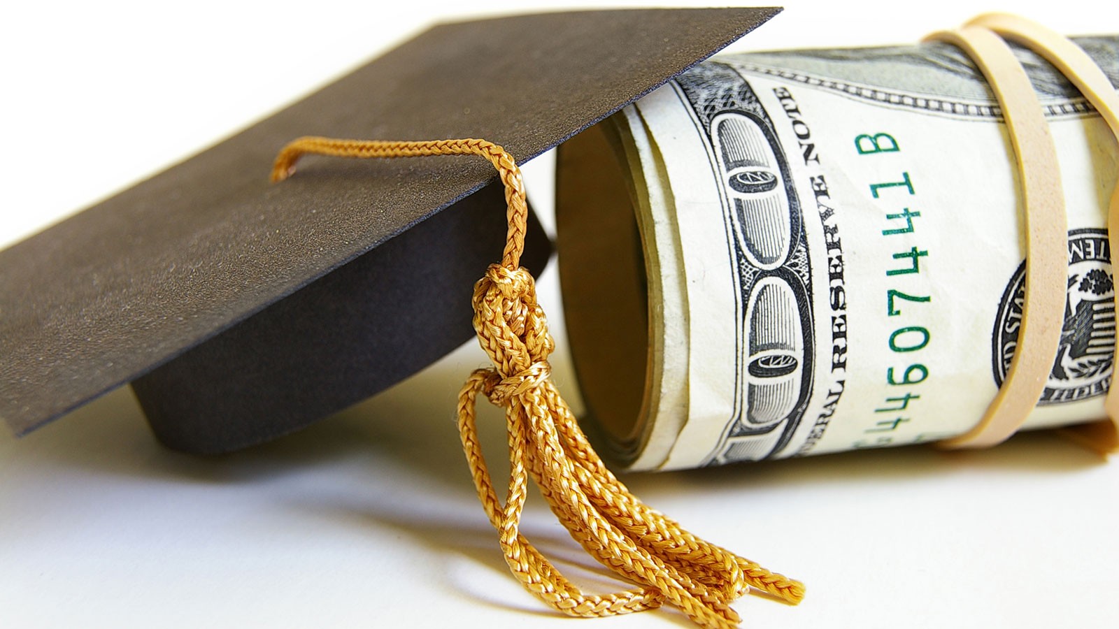 A tiny replica graduation cap rests against a roll of hundred-dollar bills.