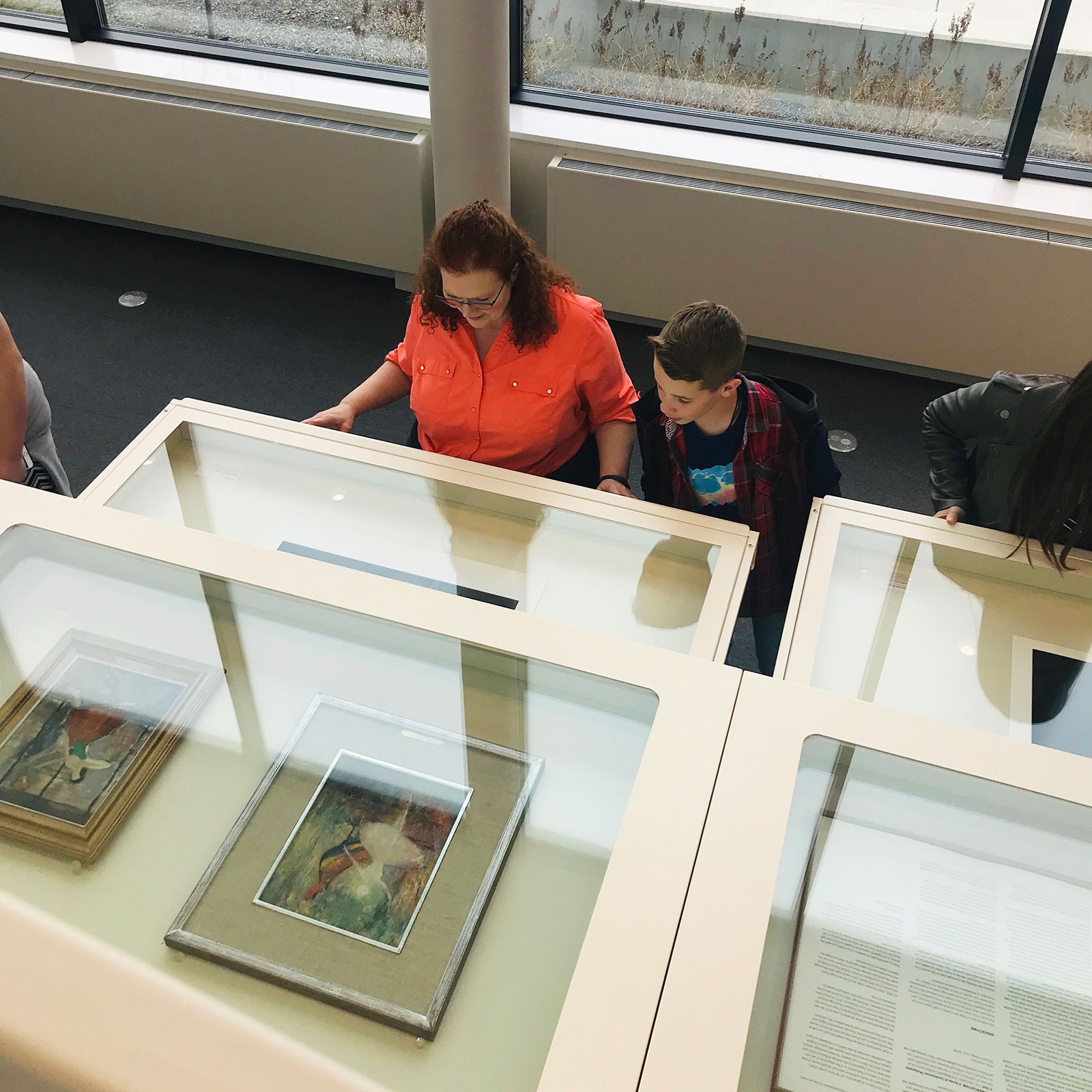 A woman and a boy study framed paintings in a museum display case.